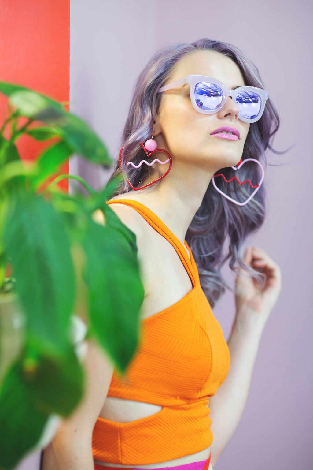 Woman wearing heart-shaped earrings and colorful sunglasses with a plant in the foreground.