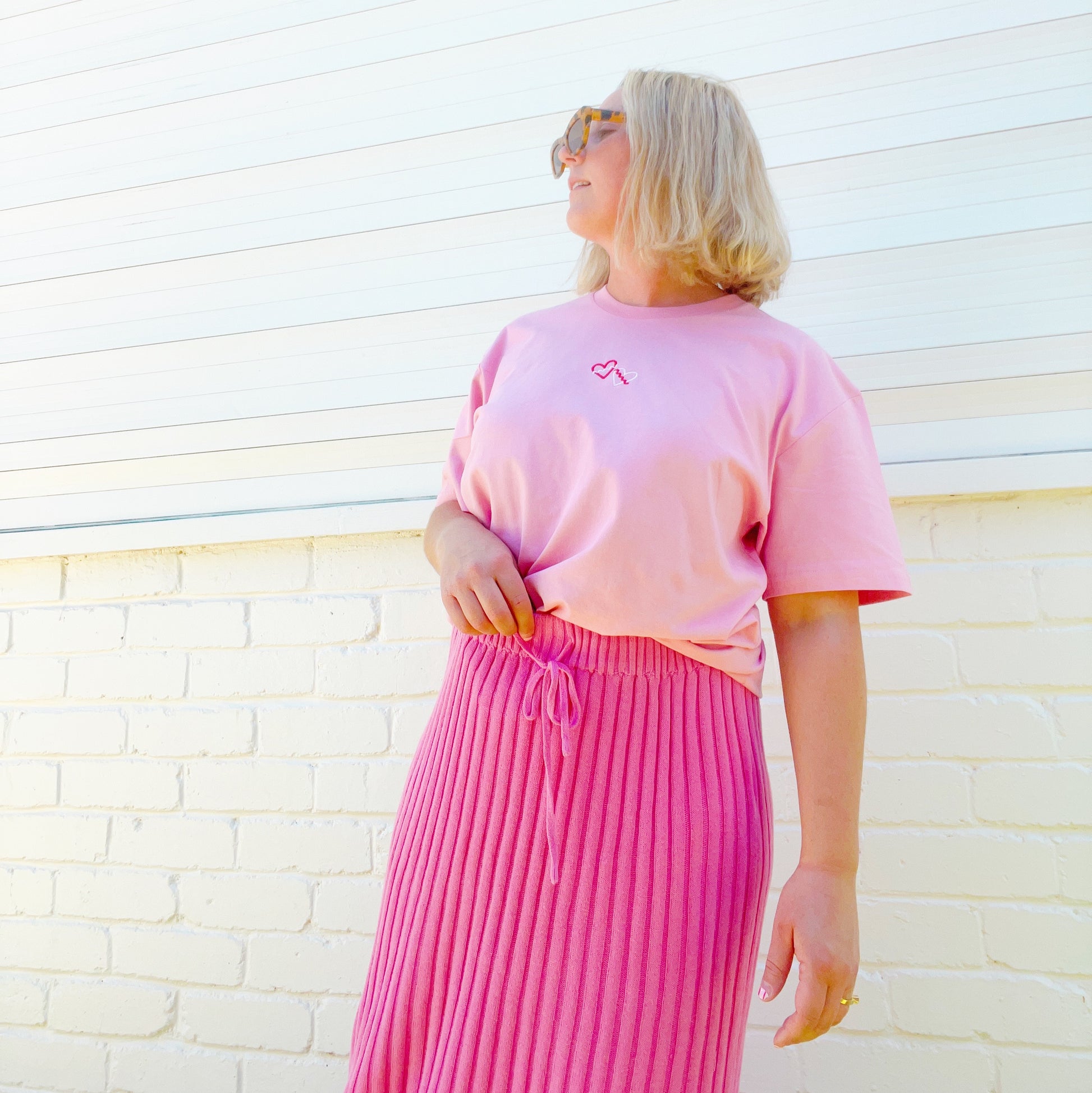 Person wearing a pink t-shirt and pleated pink skirt against a light brick wall.