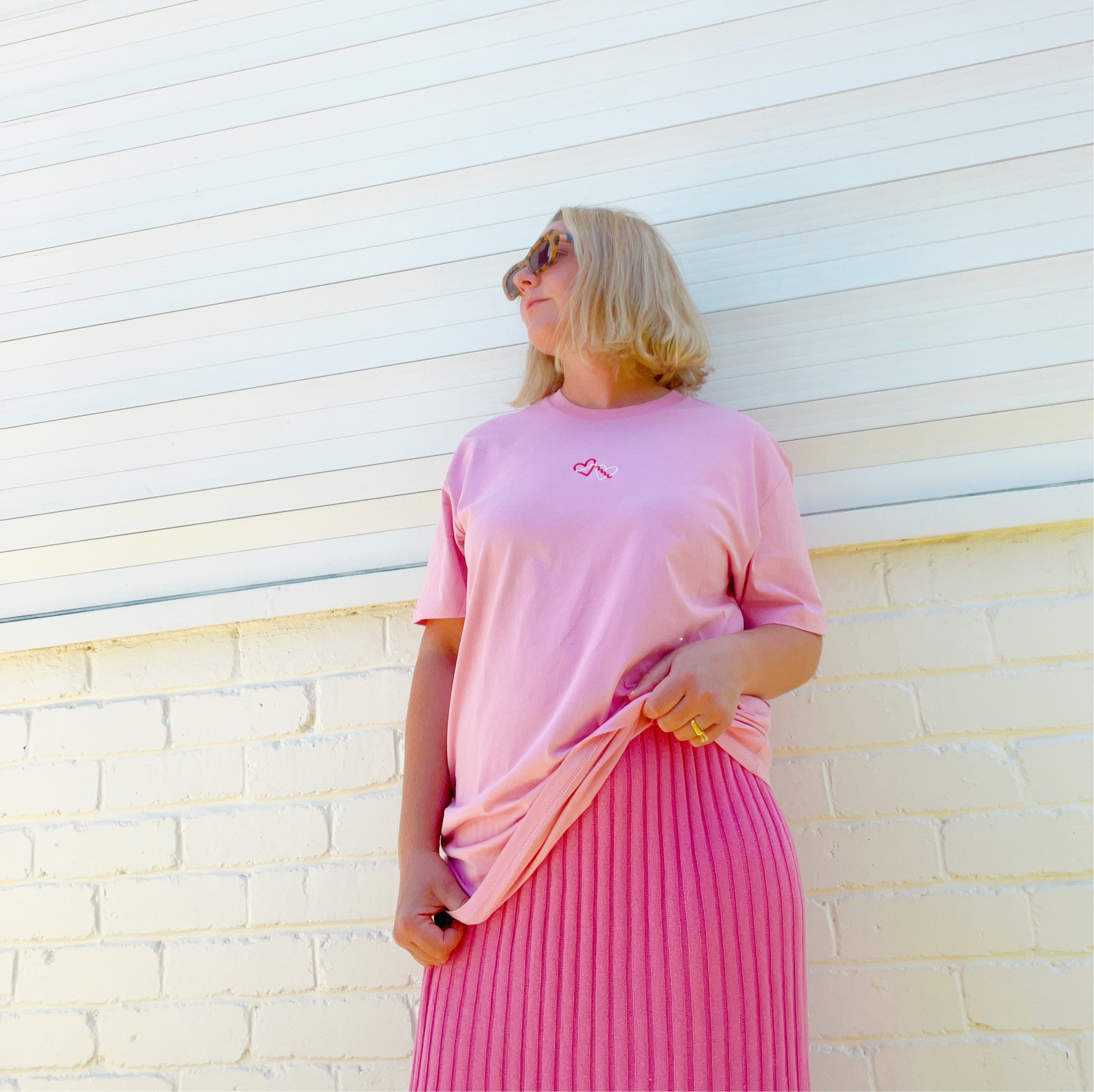 Woman wearing a pink t-shirt and pleated pink skirt against a light-colored wall.