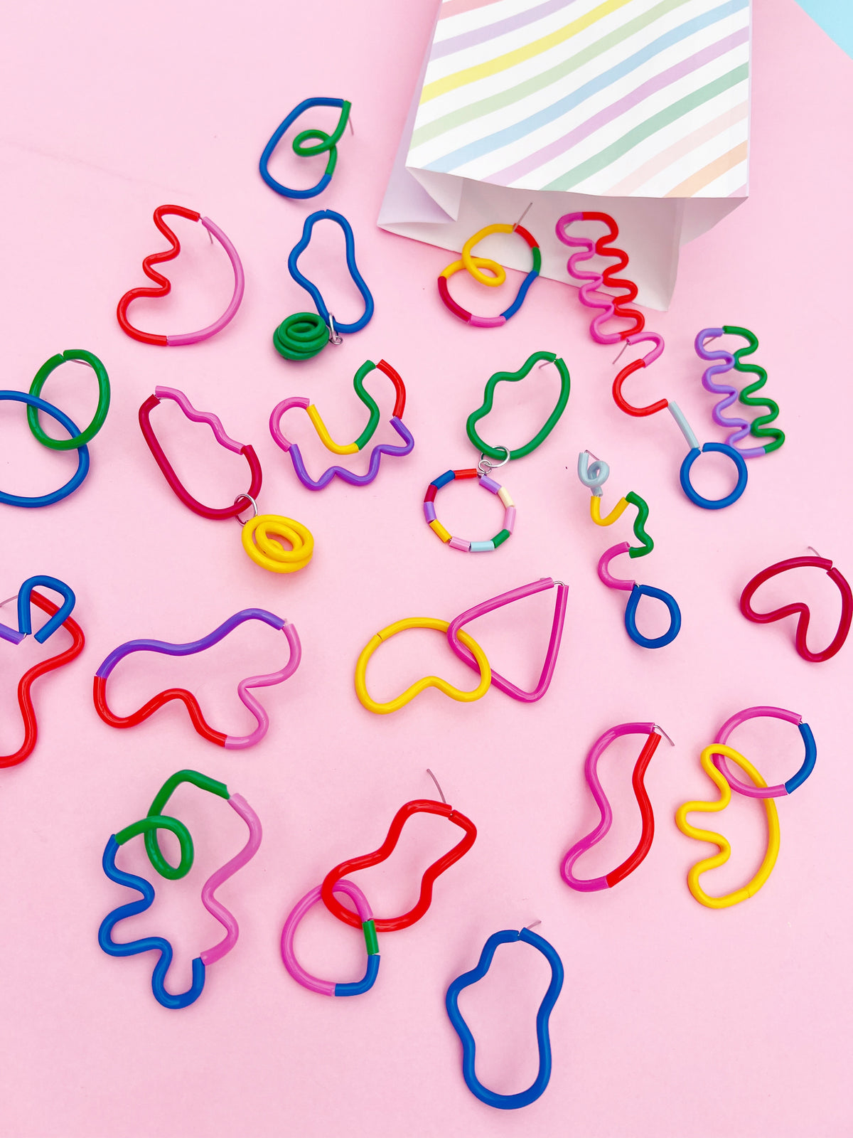 A collection of colorful, novelty, lolly like stud statement earrings in various organic shapes scattered on a light pink background, with a striped paper bag visible in the top right corner.