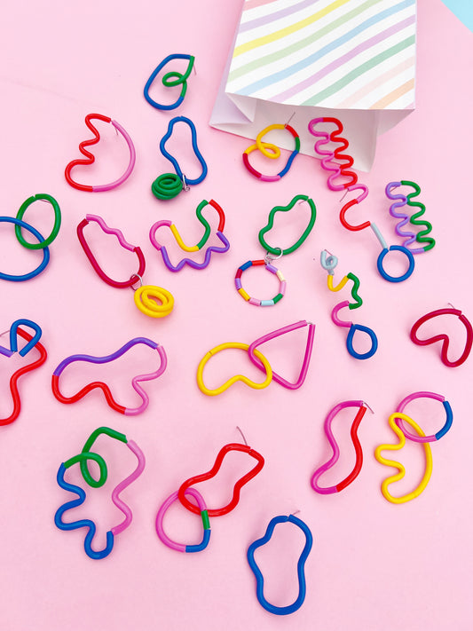 A collection of colorful, novelty, lolly like stud statement earrings in various organic shapes scattered on a light pink background, with a striped paper bag visible in the top right corner.