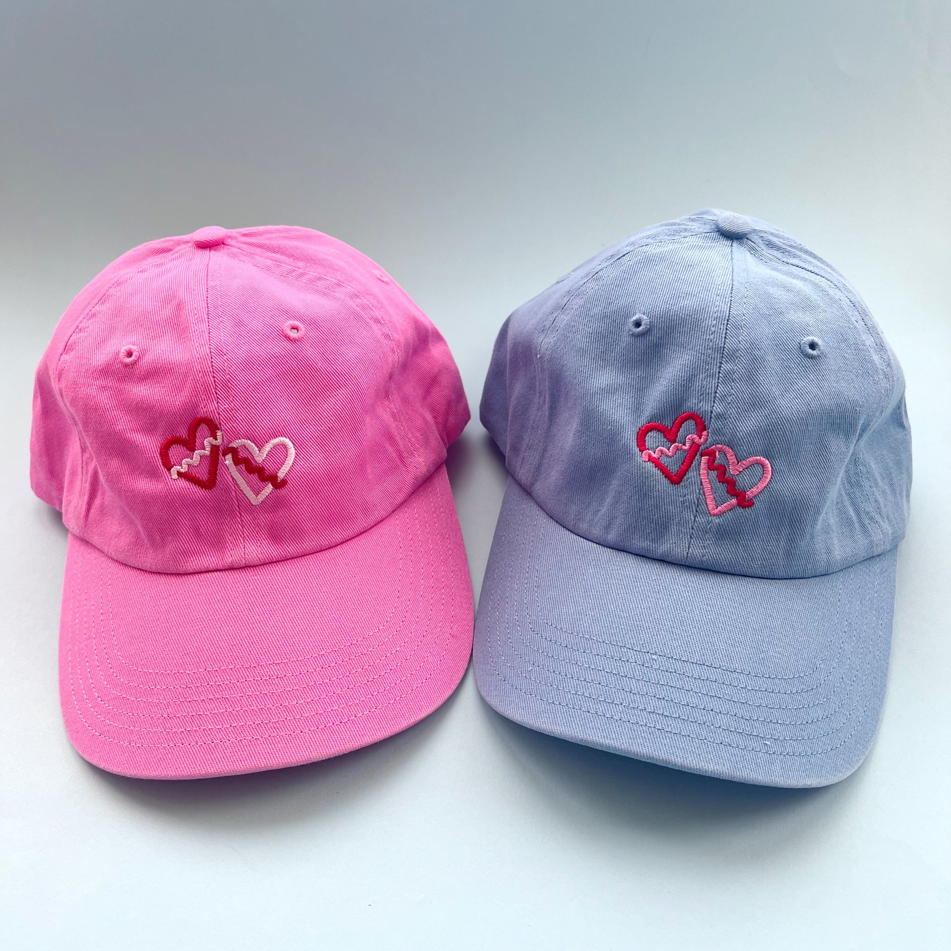 Pink and blue baseball caps with heart designs on a light gray background