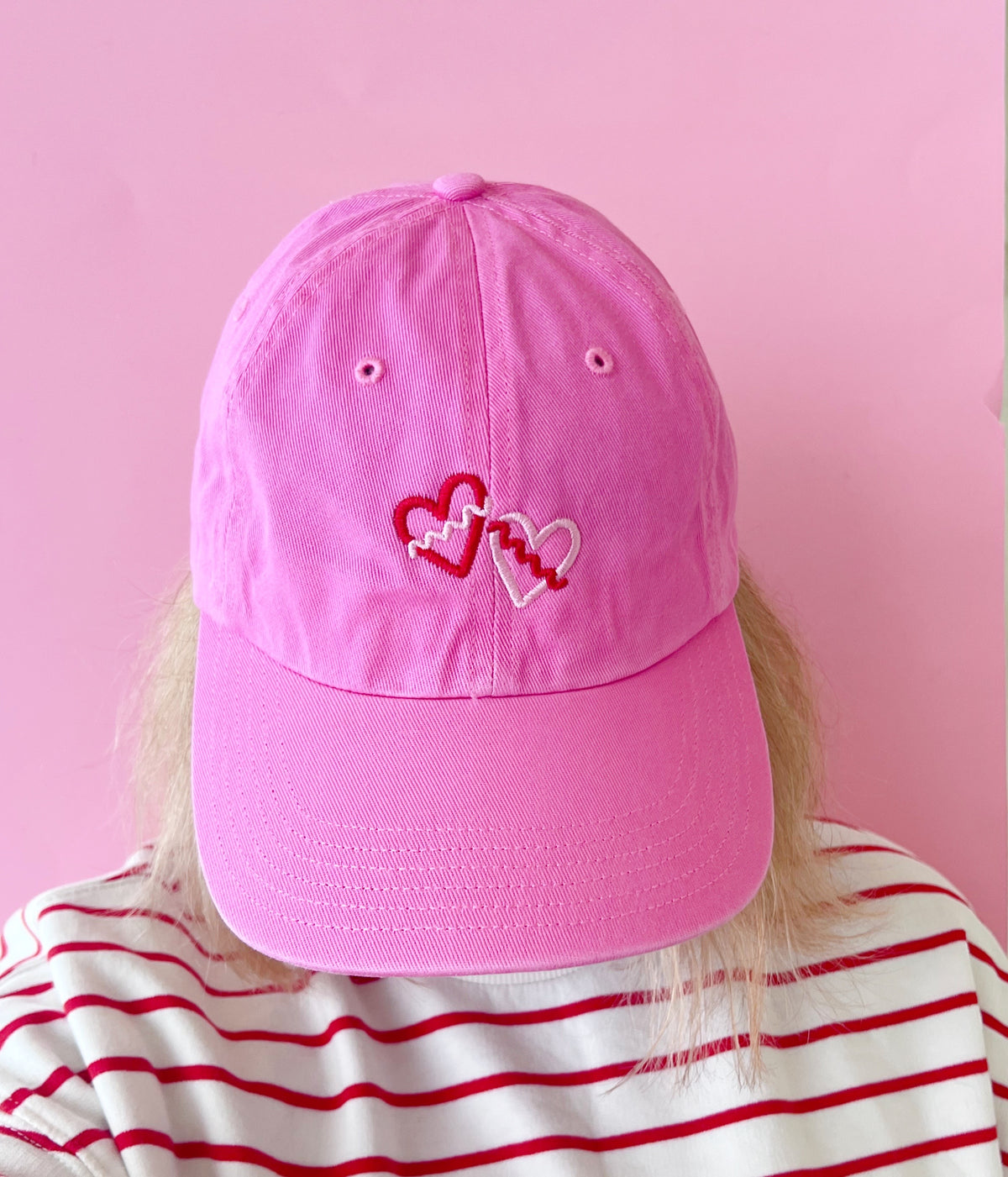 Pink cap with heart designs worn by a person against a pink background