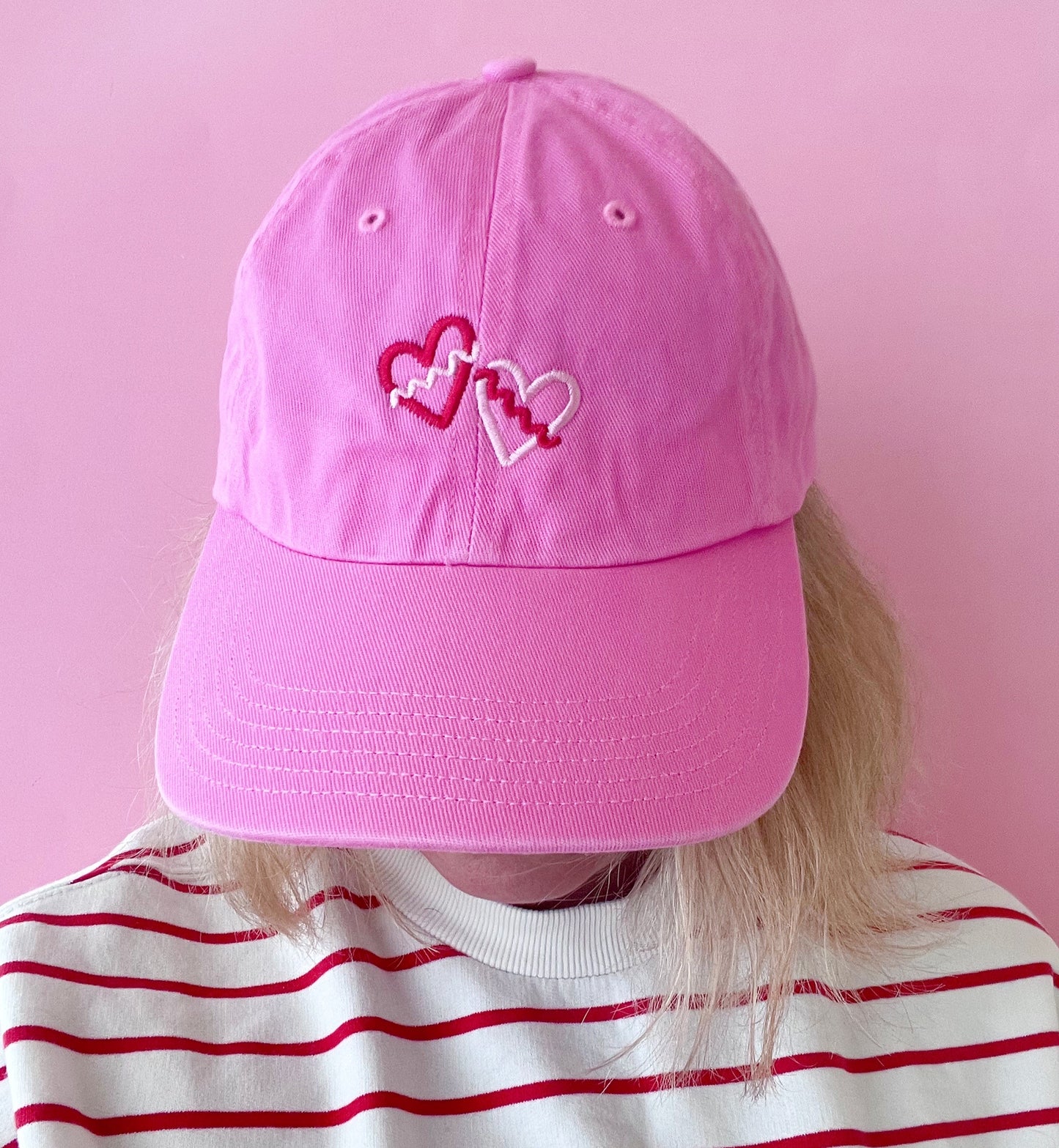 Pink cap with heart designs worn by a person against a pink background