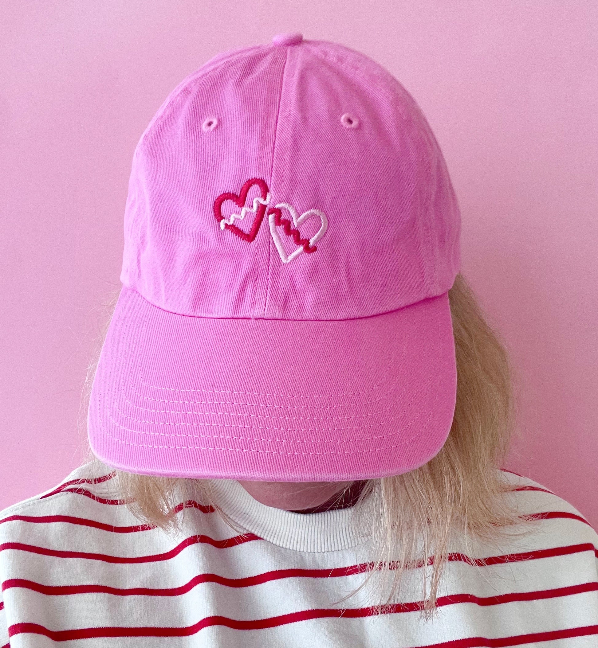 Pink cap with heart designs worn by a person against a pink background