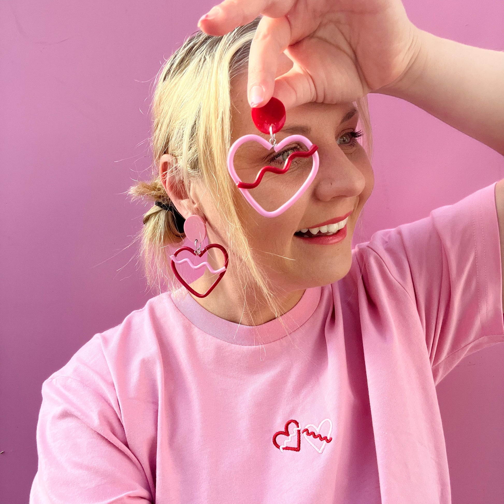 Person wearing pink heart-shaped earrings and a pink shirt with a heart design against a pink background