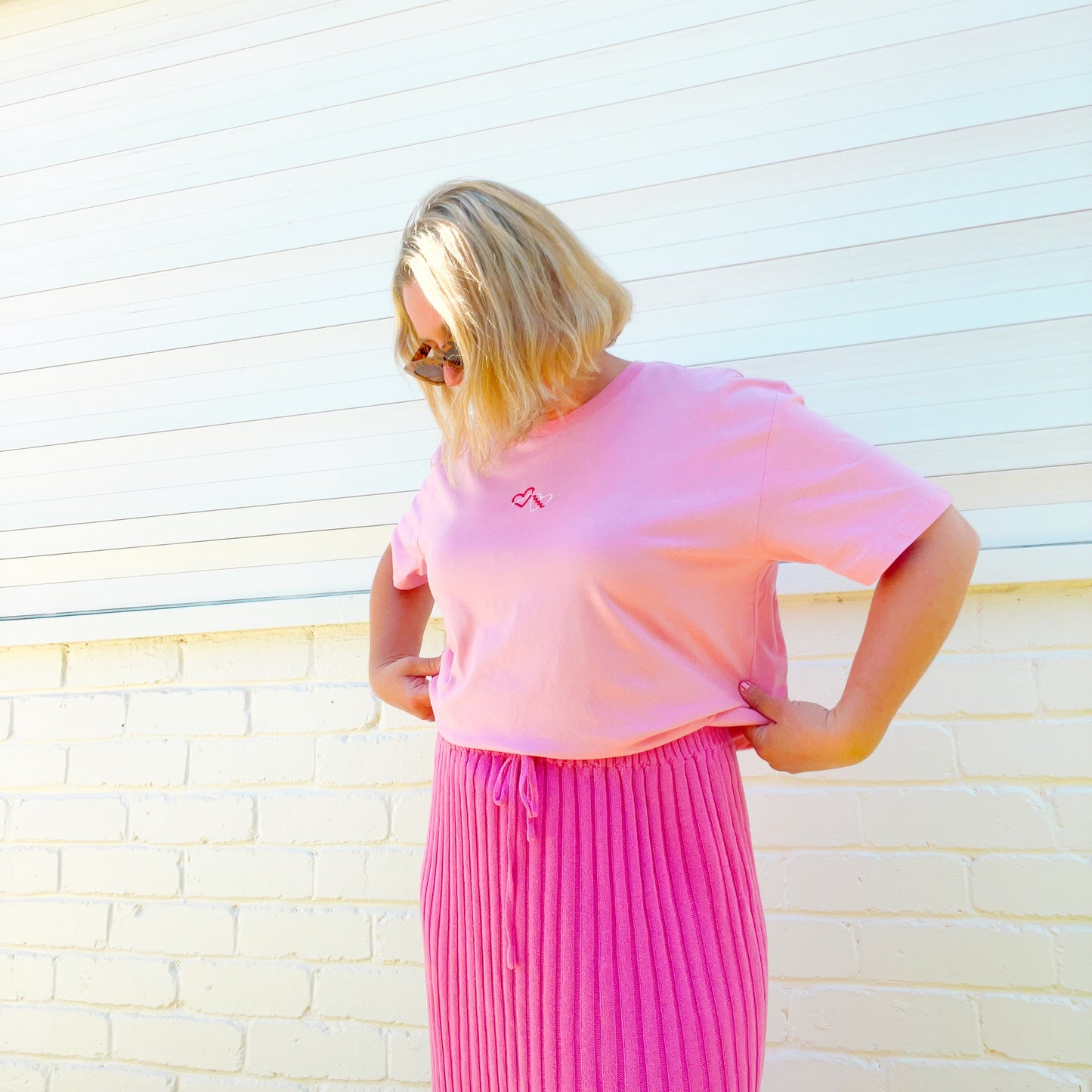 Woman wearing a pink tshirt with a heart embroidered logo against a light-colored wall.